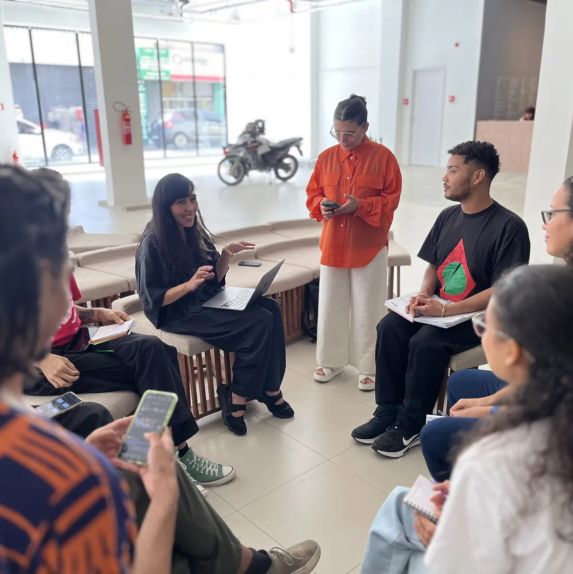 Participante de preto apresenta algo no notebook ao grupo; facilitadora de laranja acompanha a conversa em espaço invidraçado.