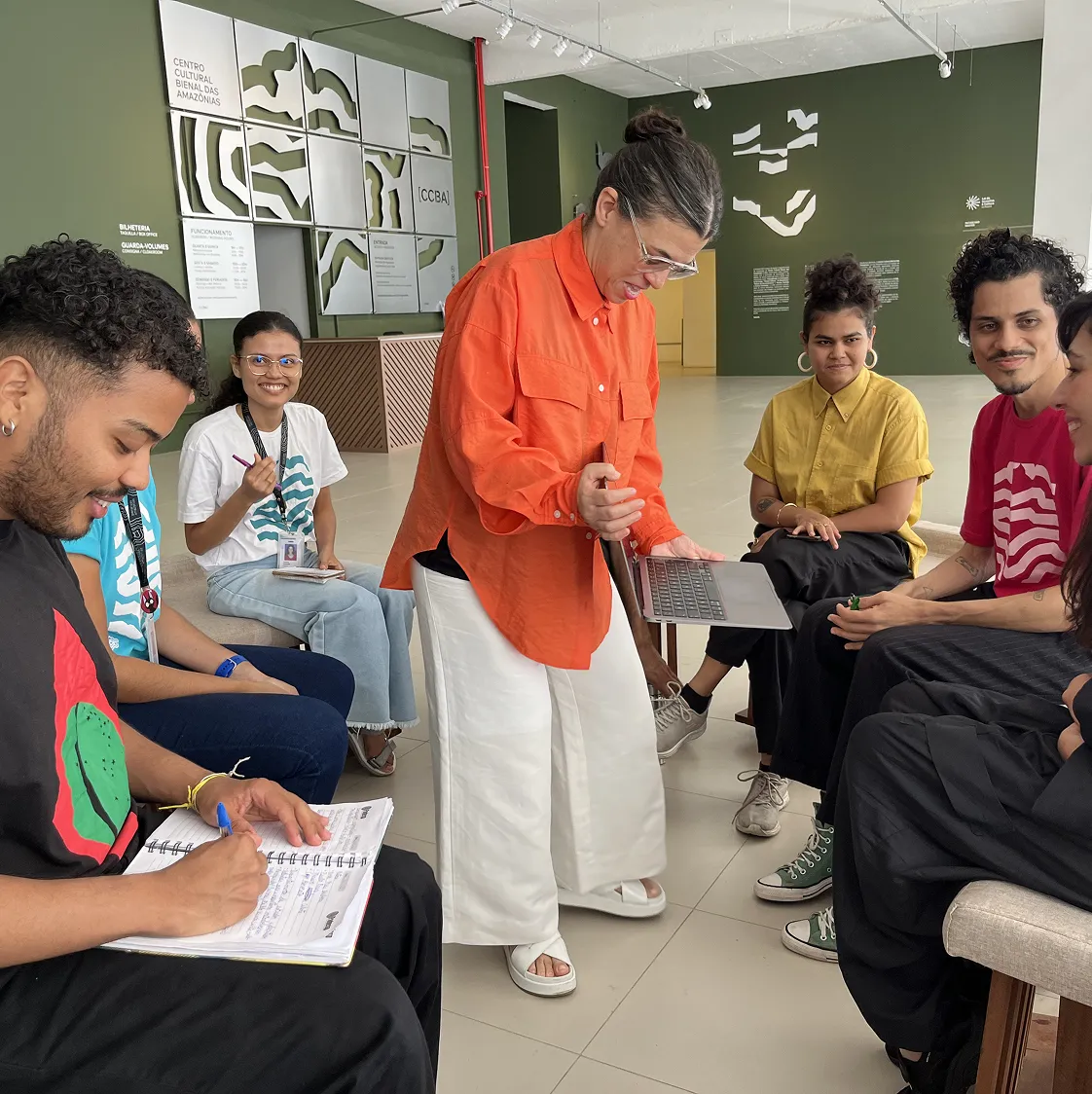 Facilitadora de camisa laranja mostra um notebook ao grupo sentado, durante atividade no hall do Centro Cultural Bienal das Amazônias.